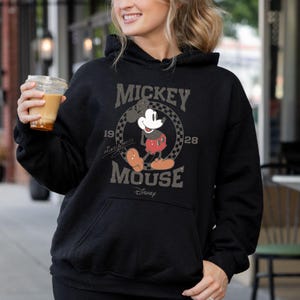May include: Black hooded sweatshirt featuring a distressed graphic of Mickey Mouse. The design includes the words "Mickey Mouse" and the years "1928" and "19". The Disney logo is at the bottom of the graphic.