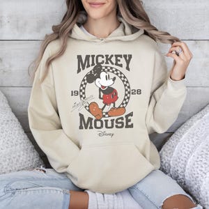 May include: A beige hoodie featuring a distressed graphic of Mickey Mouse. The design includes the words "Mickey Mouse" and "Disney" along with the year "1928". The hoodie has a front pocket and a drawstring hood.