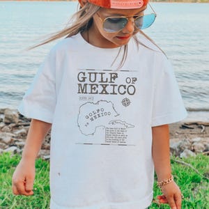 May include: White t-shirt with a brown graphic of a map of the Gulf of Mexico. The text "Gulf of Mexico" and "Est. 1972" is printed on the shirt. The graphic also includes the text "Golfo de Mexico" and a compass.