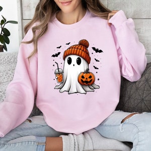 Op de afbeelding: Een lichtroze sweatshirt met een cartoongeest met een oranje muts. De geest houdt een pompoen en een drankje met een rietje vast. Zwarte vleermuizen en sterren staan op de achtergrond. Een leuk Halloween-ontwerp.