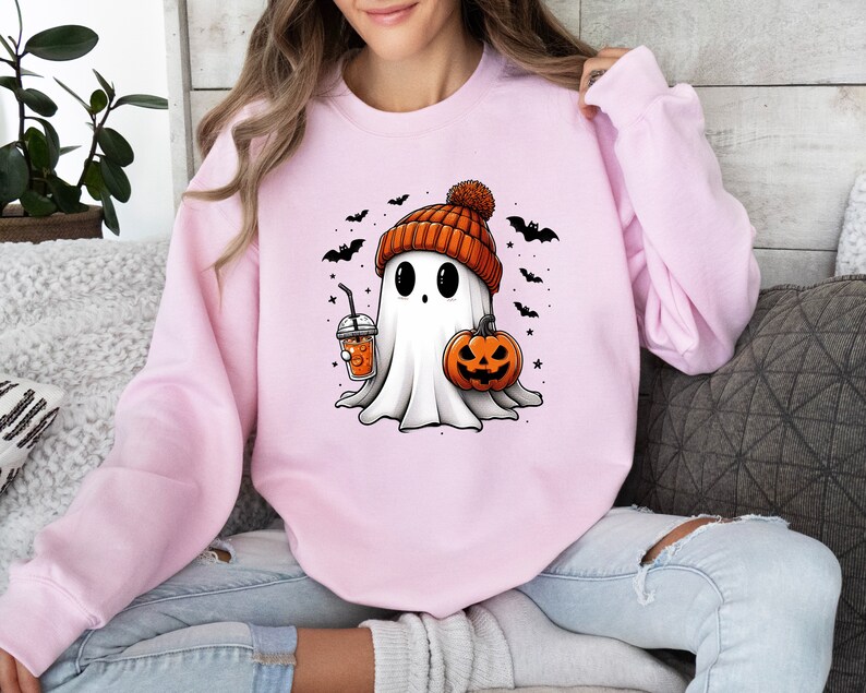 Op de afbeelding: Een lichtroze sweatshirt met een cartoongeest met een oranje muts. De geest houdt een pompoen en een drankje met een rietje vast. Zwarte vleermuizen en sterren staan op de achtergrond. Een leuk Halloween-ontwerp.