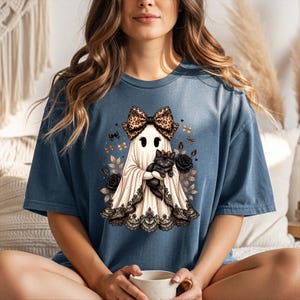 K&ouml;nnte beinhalten: Ein blaues T-Shirt mit einer Grafik eines wei&szlig;en Geistes, der eine Schleife mit Leopardenmuster tr&auml;gt und ein schwarzes K&auml;tzchen h&auml;lt. Der Geist ist am Saum mit schwarzer Spitze verziert und von schwarzen Rosen und kleinen goldenen Schmetterlingen umgeben.