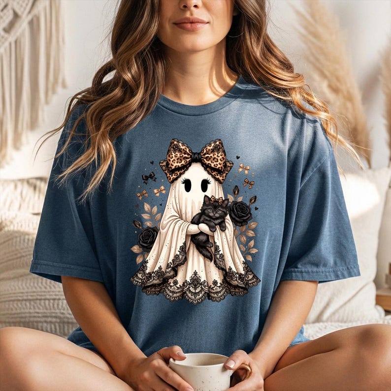 K&ouml;nnte beinhalten: Ein blaues T-Shirt mit einer Grafik eines wei&szlig;en Geistes, der eine Schleife mit Leopardenmuster tr&auml;gt und ein schwarzes K&auml;tzchen h&auml;lt. Der Geist ist am Saum mit schwarzer Spitze verziert und von schwarzen Rosen und kleinen goldenen Schmetterlingen umgeben.