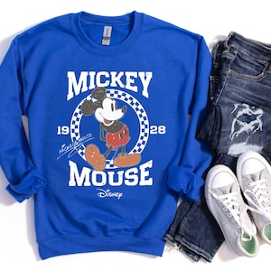 Op de afbeelding: Koningsblauw sweatshirt met een Mickey Mouse-afbeelding. Het ontwerp bevat de tekst "Mickey Mouse" en "1928" in het wit, met Mickey in zijn klassieke pose. Het sweatshirt wordt gecombineerd met een blauwe jeans en grijze sneakers.