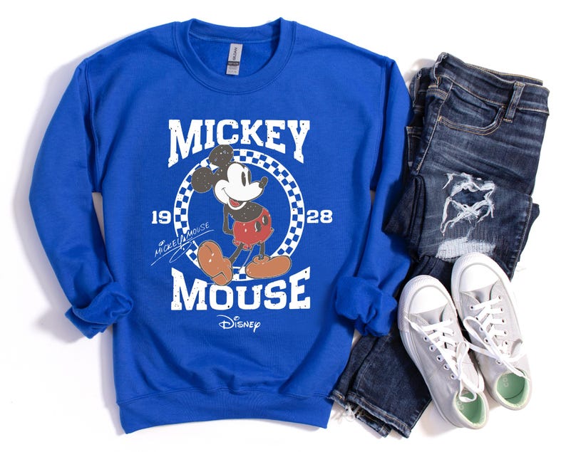 Op de afbeelding: Koningsblauw sweatshirt met een Mickey Mouse-afbeelding. Het ontwerp bevat de tekst "Mickey Mouse" en "1928" in het wit, met Mickey in zijn klassieke pose. Het sweatshirt wordt gecombineerd met een blauwe jeans en grijze sneakers.