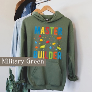 May include: Military green hoodie with the words "MASTER BUILDER" in colorful block letters. The design includes scattered colorful block shapes and stars. The hoodie has a front pocket and a hood.