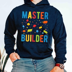 May include: Navy blue hooded sweatshirt with the words "MASTER BUILDER" in colorful block letters. The design includes scattered toy building blocks in various colors, such as red, yellow, and blue.