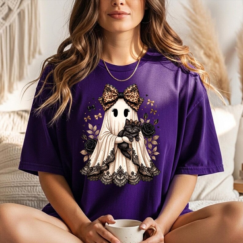 K&ouml;nnte beinhalten: Ein violettes T-Shirt mit einer Grafik eines wei&szlig;en Geistes, der eine Schleife mit Leopardenmuster tr&auml;gt und ein schwarzes K&auml;tzchen h&auml;lt. Der Geist ist am Saum mit schwarzer Spitze verziert und von schwarzen Rosen, kleinen goldenen Schleifen und Bl&auml;ttern umgeben.