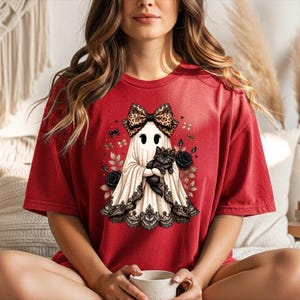 K&ouml;nnte beinhalten: Ein rotes Kurzarm-T-Shirt mit einer Grafik eines wei&szlig;en Geistes, der eine gro&szlig;e Schleife mit Leopardenmuster tr&auml;gt und ein schwarzes K&auml;tzchen h&auml;lt. Der Geist ist mit schwarzer Spitzenborte am Saum verziert und von schwarzen Rosen und kleinen goldenen Schleifen umgeben.
