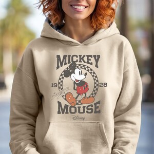 May include: A tan hoodie featuring a distressed graphic of Mickey Mouse. The design includes the words "Mickey Mouse" and the year "1928". The Disney logo is at the bottom. The hoodie has a drawstring hood and a front pocket.