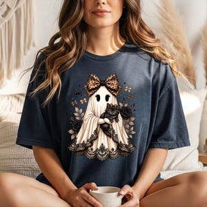 K&ouml;nnte beinhalten: Ein dunkel anthrazitfarbenes T-Shirt mit einer Grafik eines wei&szlig;en Geistes, der eine Schleife mit Leopardenmuster tr&auml;gt und eine schwarze Katze h&auml;lt. Der Geist ist von schwarzen Rosen und kleinen Schleifen umgeben, mit schwarzer Spitzenborte am Saum.