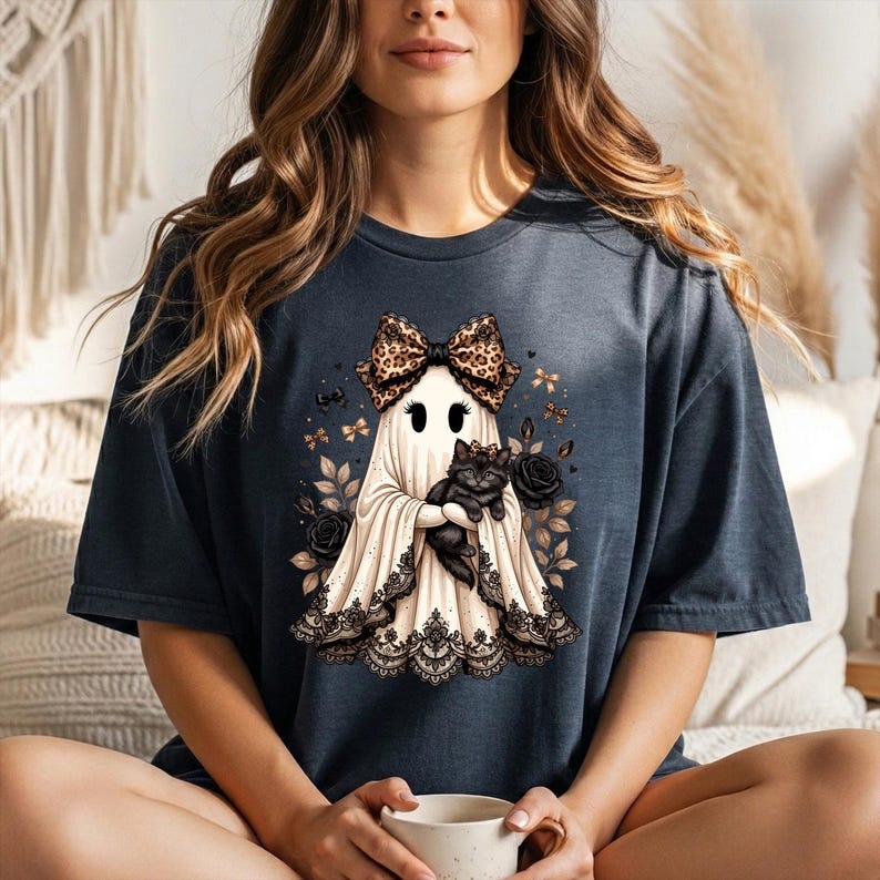 K&ouml;nnte beinhalten: Ein dunkel anthrazitfarbenes T-Shirt mit einer Grafik eines wei&szlig;en Geistes, der eine Schleife mit Leopardenmuster tr&auml;gt und eine schwarze Katze h&auml;lt. Der Geist ist von schwarzen Rosen und kleinen Schleifen umgeben, mit schwarzer Spitzenborte am Saum.