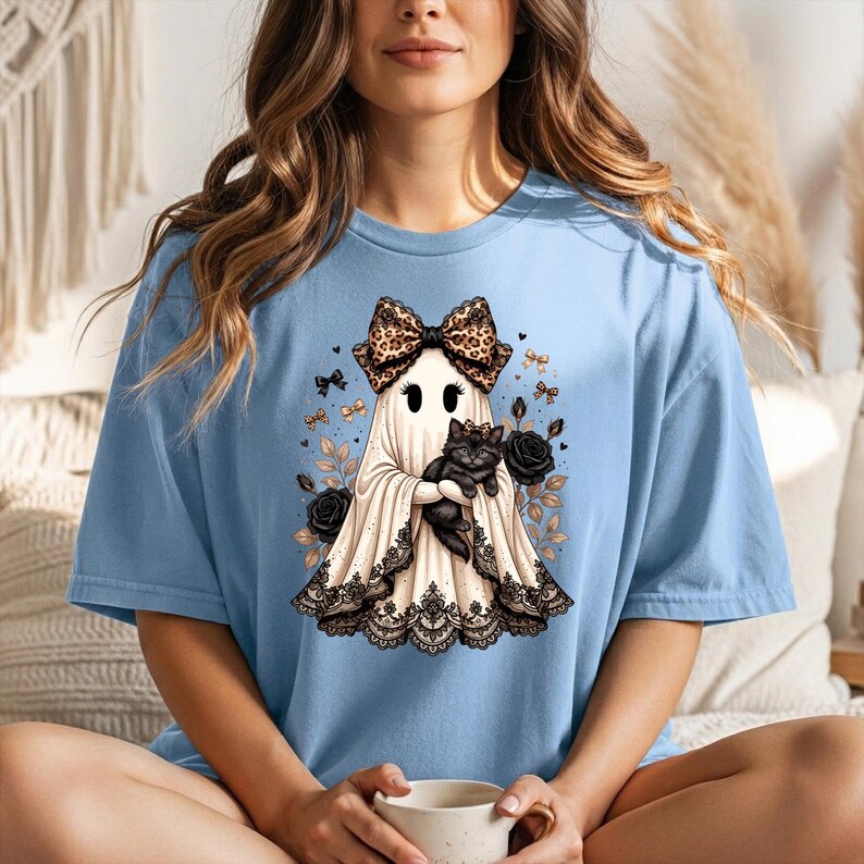 K&ouml;nnte beinhalten: Ein hellblaues T-Shirt mit einer Grafik eines Geistes, der eine Schleife mit Leopardenmuster tr&auml;gt und eine schwarze Katze h&auml;lt. Der Geist ist am Saum mit schwarzer Spitze verziert und von schwarzen Rosen, kleinen goldenen Schleifen und Zweigen umgeben.