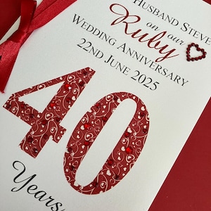 May include: A white card with red text and embellishments, celebrating a 40th wedding anniversary. The card reads "To My Husband Steve on our Ruby Wedding Anniversary 22nd June 2025". The number "40" is a focal point.