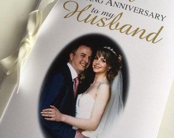 May include: A white wedding anniversary card with the text "Happy First Wedding Anniversary to my Husband" in gold script. The card features a photo of a smiling couple and is adorned with a cream-colored ribbon and small crystal embellishments.