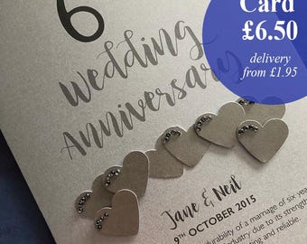 May include: A silver-colored wedding anniversary card with the text "on our 6th Wedding Anniversary". The card features heart-shaped embellishments with small, dark accents. The card also includes the names "Jane & Neil" and the date "9th October 2015".
