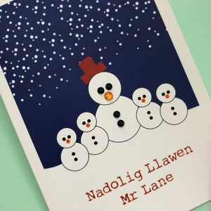 May include: A Christmas card featuring a snowman family design. The card has a dark blue background with white dots, resembling falling snow. The snowman has black gem eyes, a gem nose, and a brown hat. The text "Nadolig Llawen Mr Lane" is printed in brown.