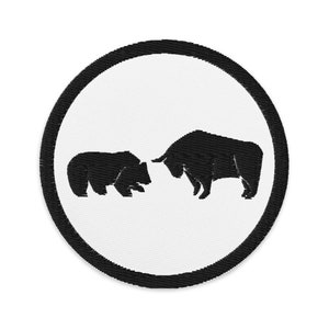 Puede incluir: Un parche bordado circular con fondo blanco y un borde grueso cosido en negro. El centro muestra la silueta negra de un oso frente a la silueta negra de un toro, posicionados horizontalmente sobre el campo circular de color blanco.