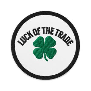 Puede incluir: Un parche redondo blanco con un borde negro. El parche tiene un trébol de cuatro hojas verde en el centro y el texto "LUCK OF THE TRADE" en un semicírculo negro sobre el trébol.