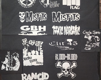 Può includere: Una collezione di toppe grafiche bianche su sfondo nero. Le toppe presentano loghi di band come "Misfits", "Black Flag" e "Rancid", insieme ad altre immagini punk rock, tra cui teschi, simboli di pace e testi come "Blood for Blood".