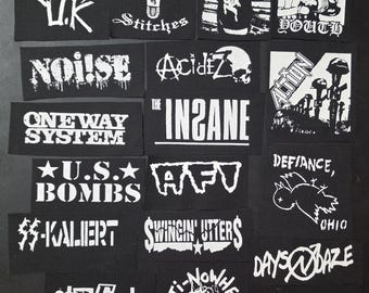 Può includere: Una collezione di toppe in bianco e nero con nomi di band punk rock e grafiche. Le toppe mostrano testi come "Chaos U.K.", "Acidiz", "U.S. Bombs" e "Swingin' Utters", insieme a varie illustrazioni.