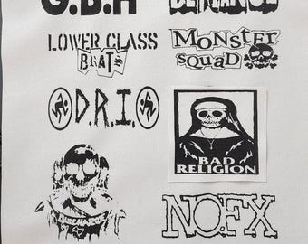Può includere: Una collezione di toppe di gruppi punk rock in bianco e nero, tra cui "NOFX", "Bad Religion", "The Casualties" e "A Global Threat". Le toppe sono su uno sfondo di tessuto bianco.