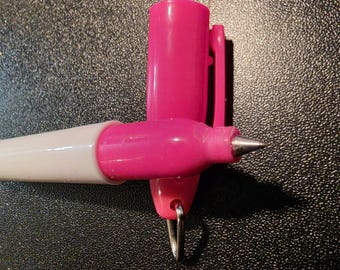Puede incluir: Un bolígrafo con cuerpo blanco y una sección de agarre de color rosa vibrante. La tapa rosa a juego está colocada en posición vertical detrás de la punta del bolígrafo, hecha de metal plateado. Un pequeño aro de metal plateado cuelga de la base.