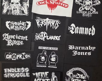 Può includere: Una collezione di toppe punk rock in bianco e nero con loghi e grafiche di band. Le toppe includono "Cock Sparrer" con un design ad ali rosse, "The Damned", "The Varukers" e "Broken Cuffs".