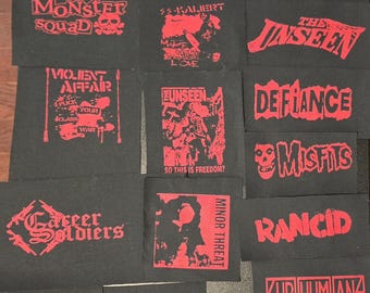 Puede incluir: Una colección de parches rectangulares de tela negra con gráficos y logotipos de bandas serigrafiados en rojo, incluyendo A Global Threat, Toxic Narcotic, Charged G.B.H, The Casualties, Monster Squad, SS-Kaliert, The Unseen, Violent Affair, Defiance, Misfits, Career Soldiers, Minor Threat, Rancid, Subhumans, Clit 45, Lower Class Brats y Agression.