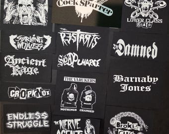 Puede incluir: Una colección de parches de tela negra con gráficos y nombres de bandas serigrafiados en blanco, incluyendo Noxious, Cock Sparrer, Lower Class Brats, Starving Wolves, The Restarts, Soap Charge, The Damned, Ancient Rage, Cropknox, The Varukers, Barnaby Jones, Endless Struggle, The Nerve Agents y Broken Cuffs.