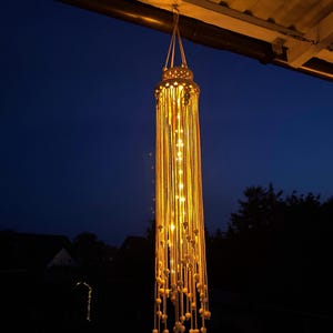 Könnte beinhalten: Eine hängende Makramee-Laterne mit warmweißen LED-Lichtern. Die Laterne hängt von einem Dach und hat ein zylindrisches Design mit langen, vertikalen Strängen und dekorativen Holzperlen. Der Hintergrund ist ein dunkelblauer Nachthimmel.