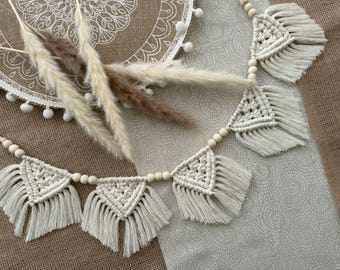 May include: Cream-colored macrame garland with wooden beads and fringed triangles. The garland is displayed with dried pampas grass and a white dream catcher with pom-poms, creating a bohemian aesthetic. The background is a neutral fabric.