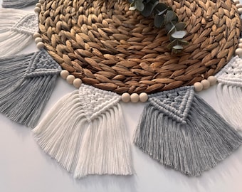 May include: A macrame wall hanging with gray and white tassels and wooden beads. The tassels are arranged in a semi-circle shape and are hanging from a woven mat.