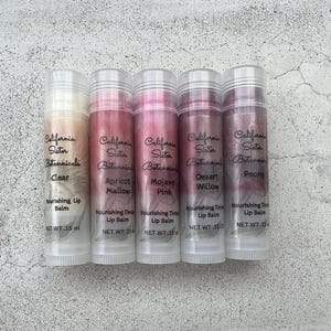 May include: Five lip balms in clear tubes with silver caps. The balms are labeled "California Sister Botanicals" and include shades like "Clear," "Apricot Mallow," and "Mojave Pink." Each tube contains 0.15 oz of product.