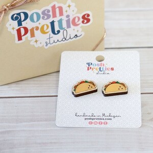May include: A pair of taco-shaped earrings on a white card, next to a beige gift box tied with a rose gold ribbon. The card and box have the "Posh Pretties studio" logo. The earrings are brown, yellow, red, and green.