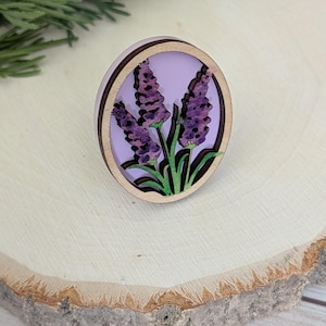 May include: A round wooden pin featuring a lavender design. The pin has a light purple background with three purple lavender flower stems and green leaves. The pin is set on a wooden surface.