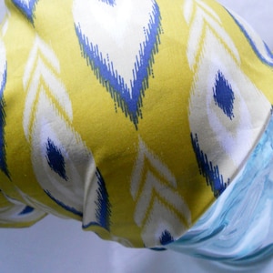 Puede incluir: Una tela amarilla con un patrón blanco y azul en forma de pluma está drapeada sobre un objeto de vidrio transparente azul claro. El diseño de la tela presenta formas repetidas en amarillo, blanco y azul. El objeto de vidrio tiene una forma curva.