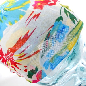 Puede incluir: Un pañuelo colorido con un estampado floral. La tela presenta un fondo blanco con diseños florales rojos, amarillos, azules y verdes vibrantes. El pañuelo está drapeado sobre una forma de vidrio transparente.