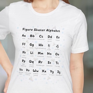 以下が含まれることがあります： 「Figure Skater Alphabet」の文字とスケート靴のイラストが描かれた白いTシャツ。シャツには、「Axel」、「Lutz」、「Salchow」などのフィギュアスケート用語とともにアルファベットが表示されています。