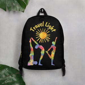 May include: Black backpack featuring the text "Travel Light" above a yellow sun graphic. Below the text are three colorful figures in yoga poses. The figures are made up of various colors including red, blue, and yellow.