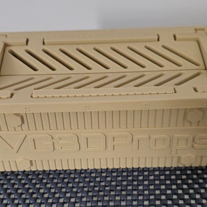 May include: Tan-colored, rectangular box with a hinged lid and diagonal vents. The box features the text "VG3DPROPS" on the front. The box is made of a rigid material and has a textured surface.