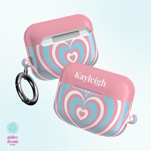 Könnte beinhalten: Zwei Schutzhüllen für kabellose Ohrhörer mit einem Muster aus konzentrischen rosa und hellblauen Herzen. Eine Hülle zeigt den Namen Kayleigh in weißer Schrift auf dem Deckel und hat einen runden Karabinerhaken aus silbernem Metall an der Seite.