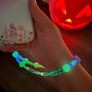 May include: Glow-in-the-dark beaded phone charm with the name "SM COREN" spelled out in white letters. The charm features large, round, glowing green and blue beads. A white phone case with floral design is visible.