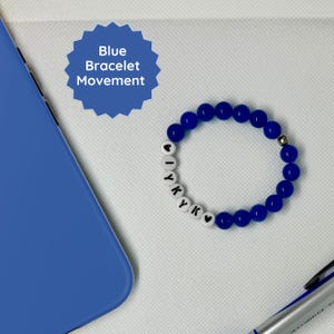 May include: Blue bracelet with white beads spelling "IYKYK" with a heart on each end. The bracelet is on a white surface with a blue phone case and a silver pen.