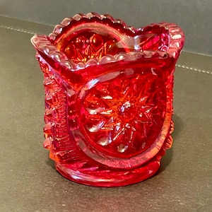 May include: A vibrant red, iridescent glass toothpick holder. The small, decorative container features a starburst pattern on the front and sides, with a scalloped rim. The glass has a textured, pressed design.
