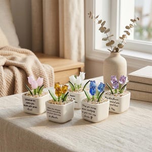 May include: Five small, square white ceramic pots each hold a glass tulip flower in shades of pink, yellow, white, blue, and purple. Each pot has a different inspirational quote. The tulips are set in small pebbles.