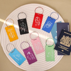 May include: A selection of luggage tags in various colours, each with a metal ring and unique design. Text includes "Frida Kahlo begins", "Go Travel Albert Einstein", and "Harper". A British passport and boarding passes are also present.