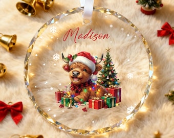 May include: A clear, faceted glass Christmas ornament with a festive design. It features a Highland cow wearing a Santa hat and scarf, surrounded by presents and a Christmas tree. The name "Madison" is written in red script.