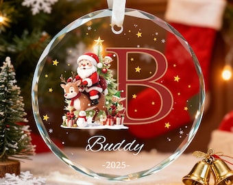 May include: A clear, round Christmas ornament with a festive design. Santa Claus rides a reindeer, with a Christmas tree and gifts. The letter "B" is in the center, with the name "Buddy" and the year "-2025-" below. A white ribbon is attached for hanging.