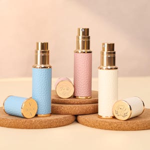 Può includere: Tre vaporizzatori di profumo in azzurro, rosa e bianco con dettagli dorati. Ogni vaporizzatore ha un esterno strutturato simile alla pelle e un ugello spray dorato. I vaporizzatori sono esposti su sottobicchieri di sughero, con tappi cilindrici abbinati.